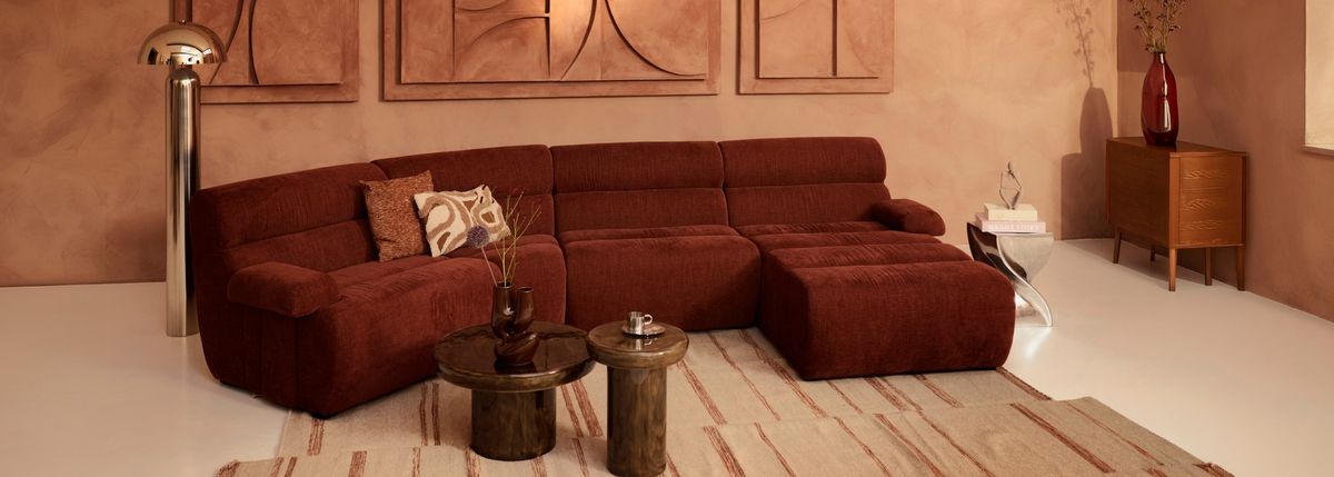 A modern living room with a rust-colored sectional sofa, decorative pillows, brown nested tables, a tall silver lamp, and abstract wall art.