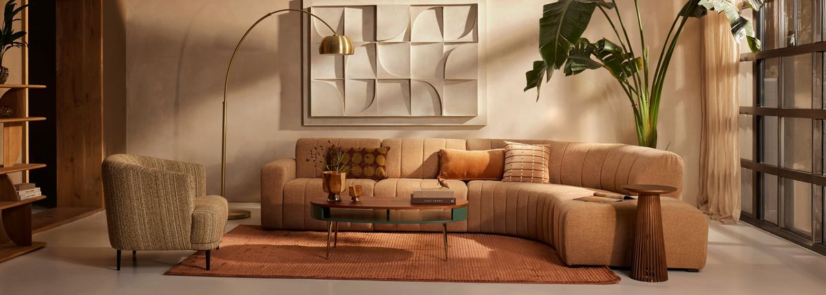 Modern living room with a beige sectional sofa, decorative pillows, wooden accents, abstract wall art, and large plants by a window with sheer curtains.