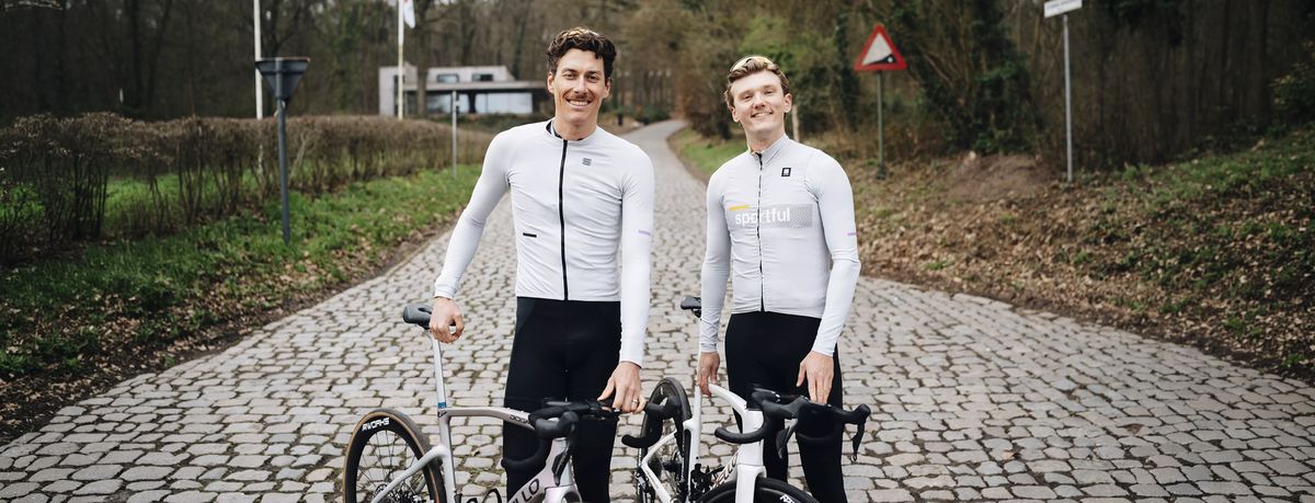 Two cyclists in white jerseys stand on a cobblestone path, holding their bikes, with greenery and road signs in the background.