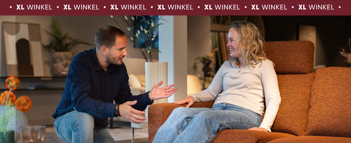 Een man en een vrouw in gesprek op een bruine bank in een moderne winkelomgeving met planten en decoratieve elementen.
