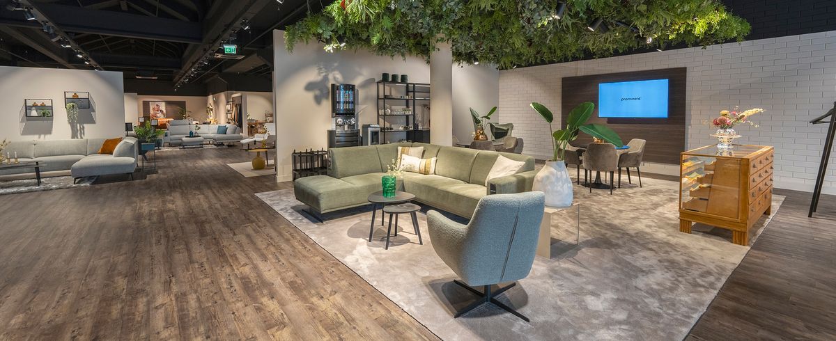 Moderne woonkamer met groene hoekbank, grijze stoel, houten vloer, planten, wandkunst en minimalistische decoratie.