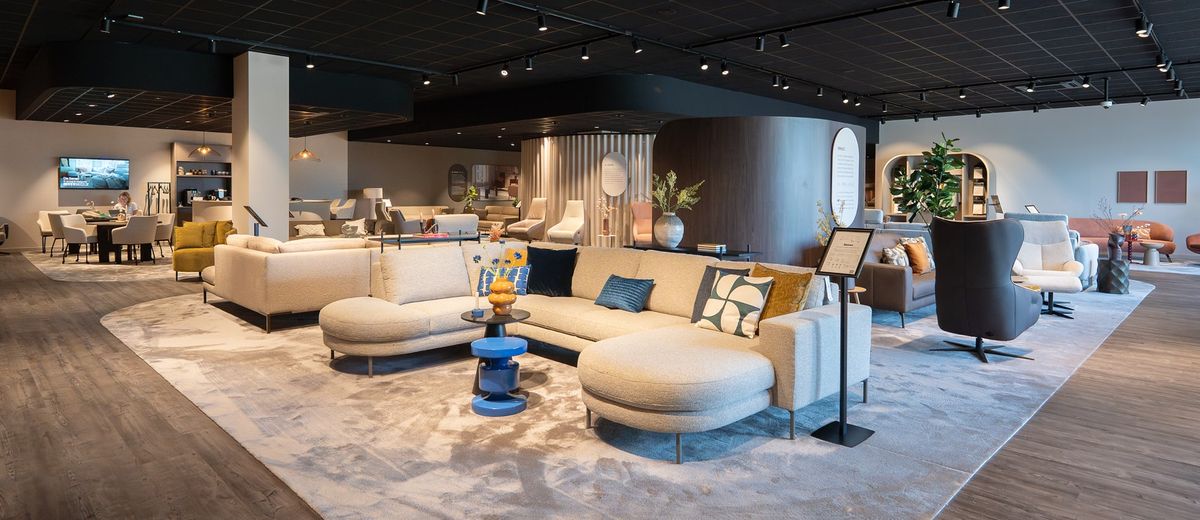 Moderne woonkameropstelling met beige hoekbank, kleurrijke kussens, blauwe bijzettafeltjes en decoratieve accessoires op een zacht tapijt.