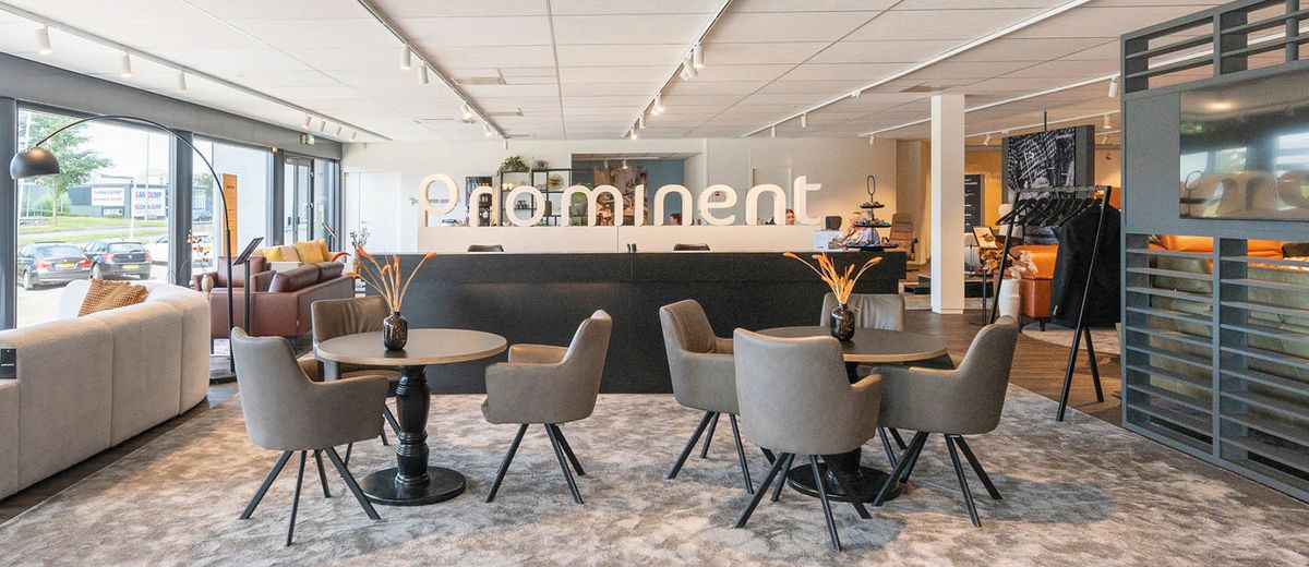 Een moderne showroom met comfortabele stoelen en tafels op een zacht tapijt, grote ramen en het woord 'prominent' op de achtergrond.
