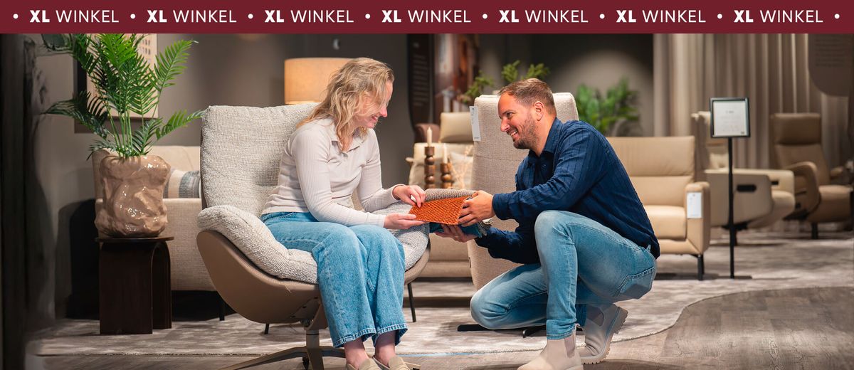 Een vrouw en een man zitten in een meubelwinkel; de vrouw zit in een stoel terwijl de man naast haar gehurkt een boek of catalogus toont.