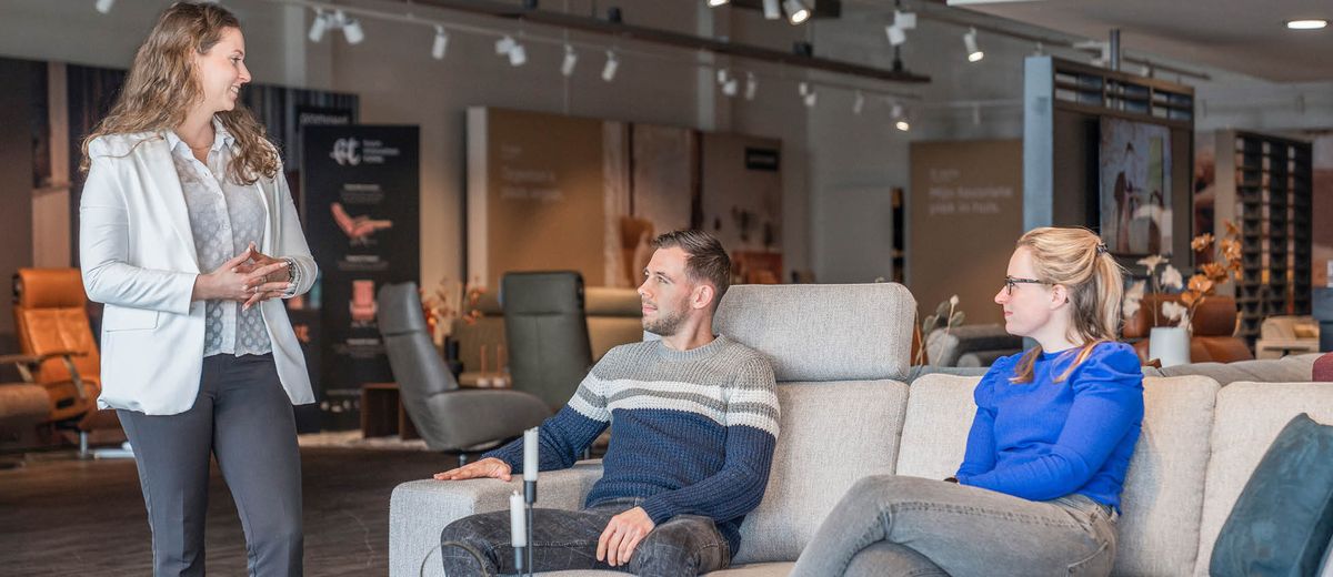 Een verkoopster toont een koppel een bank in een meubelwinkel. De man en vrouw zitten op de bank en luisteren aandachtig.