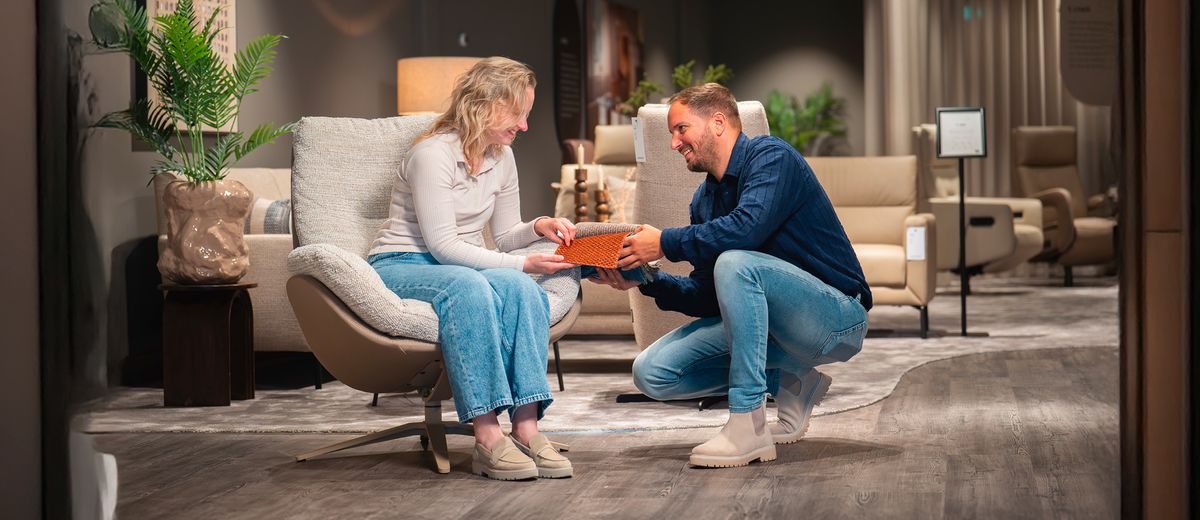 Een man en vrouw zitten in een moderne, sfeervol verlichte woonkamer. De vrouw zit in een stoel terwijl de man iets overhandigt.