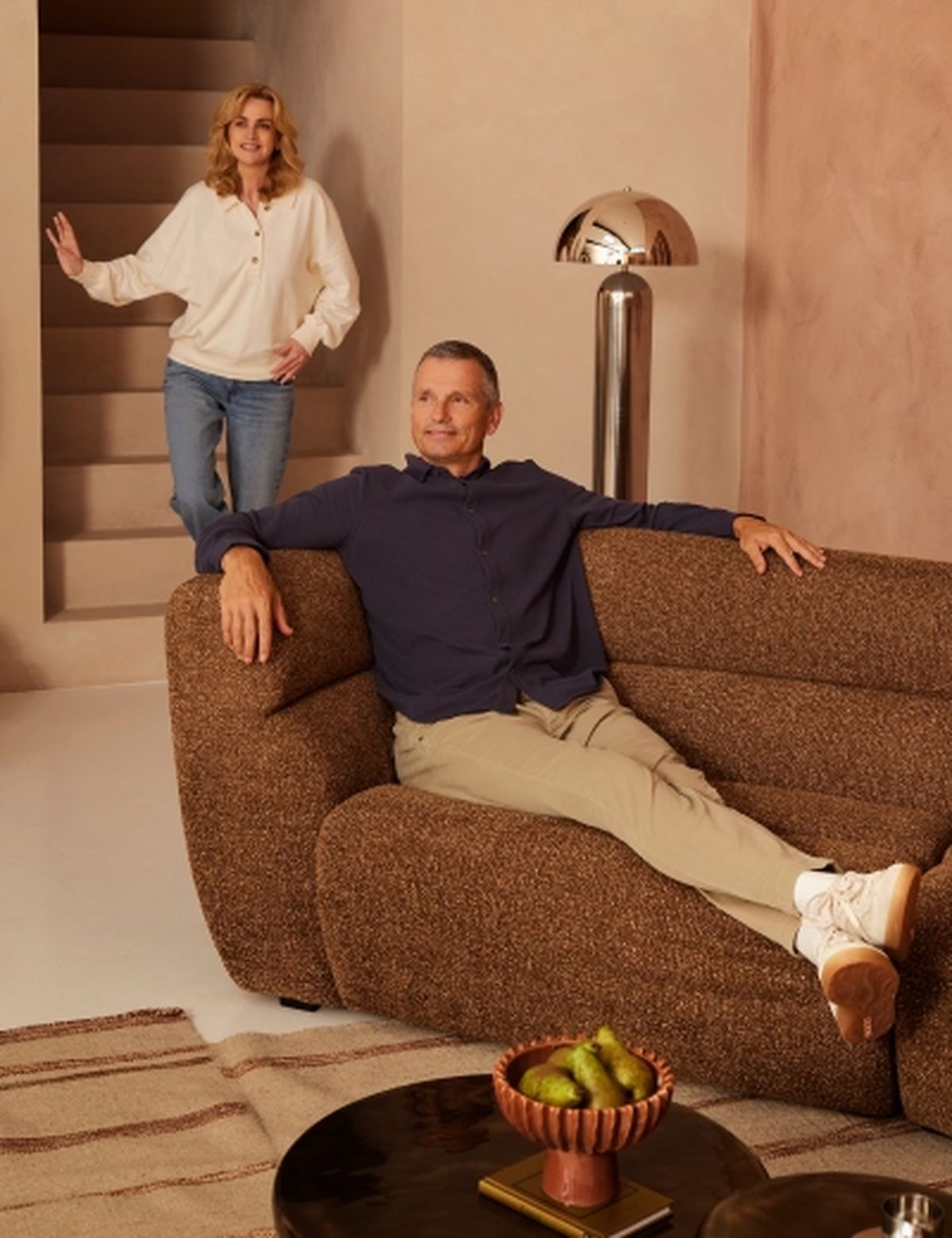 A man sits relaxed on a brown sofa, while a woman stands on stairs behind him. A round table with fruits and a lamp are nearby.