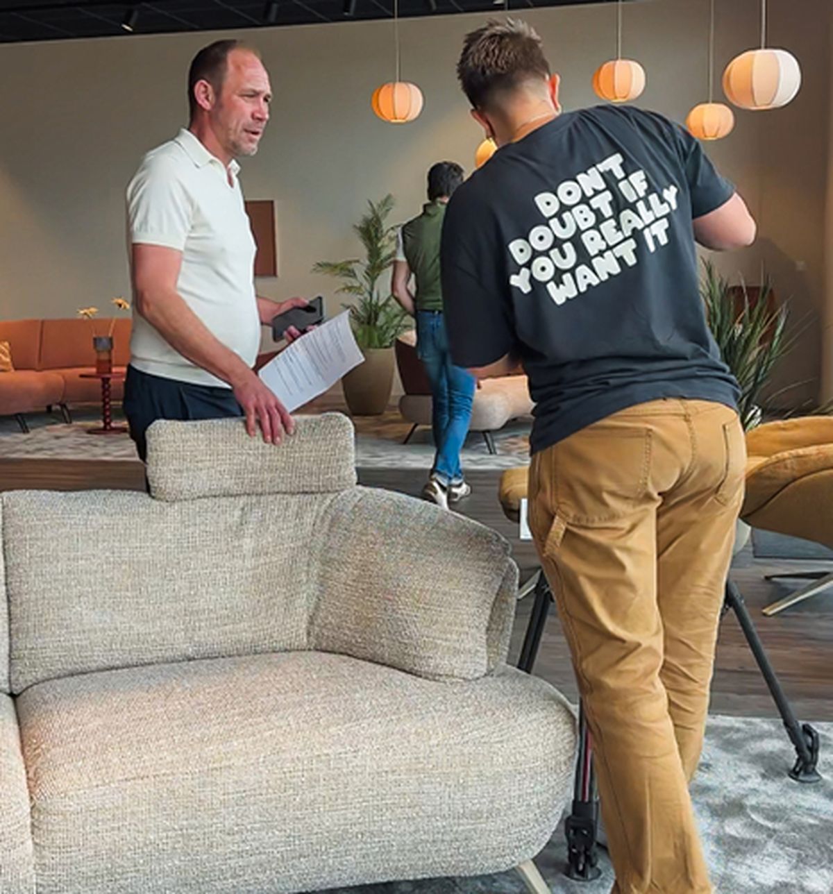 Two men in a modern living room, one carrying papers, the other adjusting a camera. Wall with lights and plants in the background.