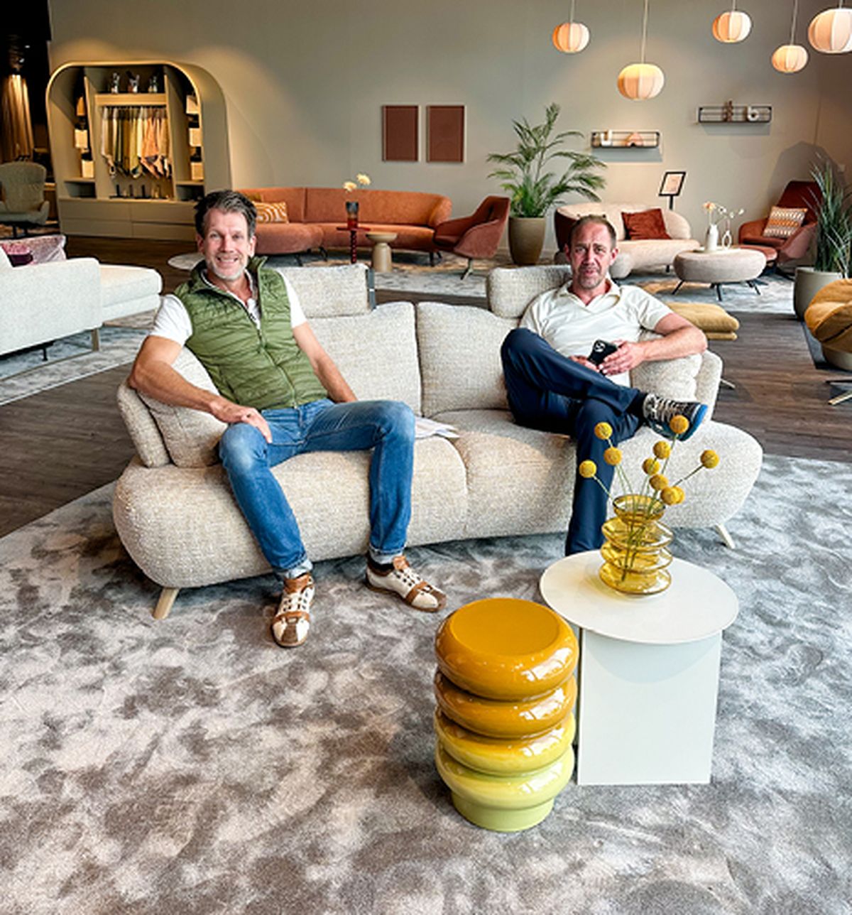 Two men sitting on a beige sofa in a modern showroom with stylish furniture, colorful cushions, and decorative lighting.