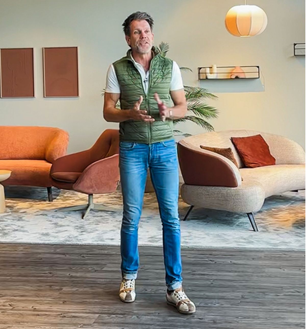 A man in a green vest and jeans is speaking in a modern, stylish room with colorful sofas, art on the wall, and ambient lighting.