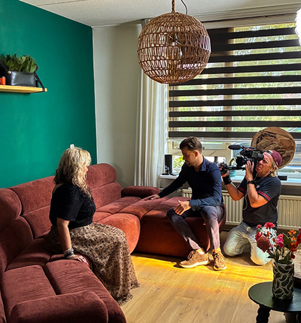 Een vrouw zit op een rode bank in een woonkamer, terwijl een cameraman haar filmt en iemand anders een lichtreflector vasthoudt.