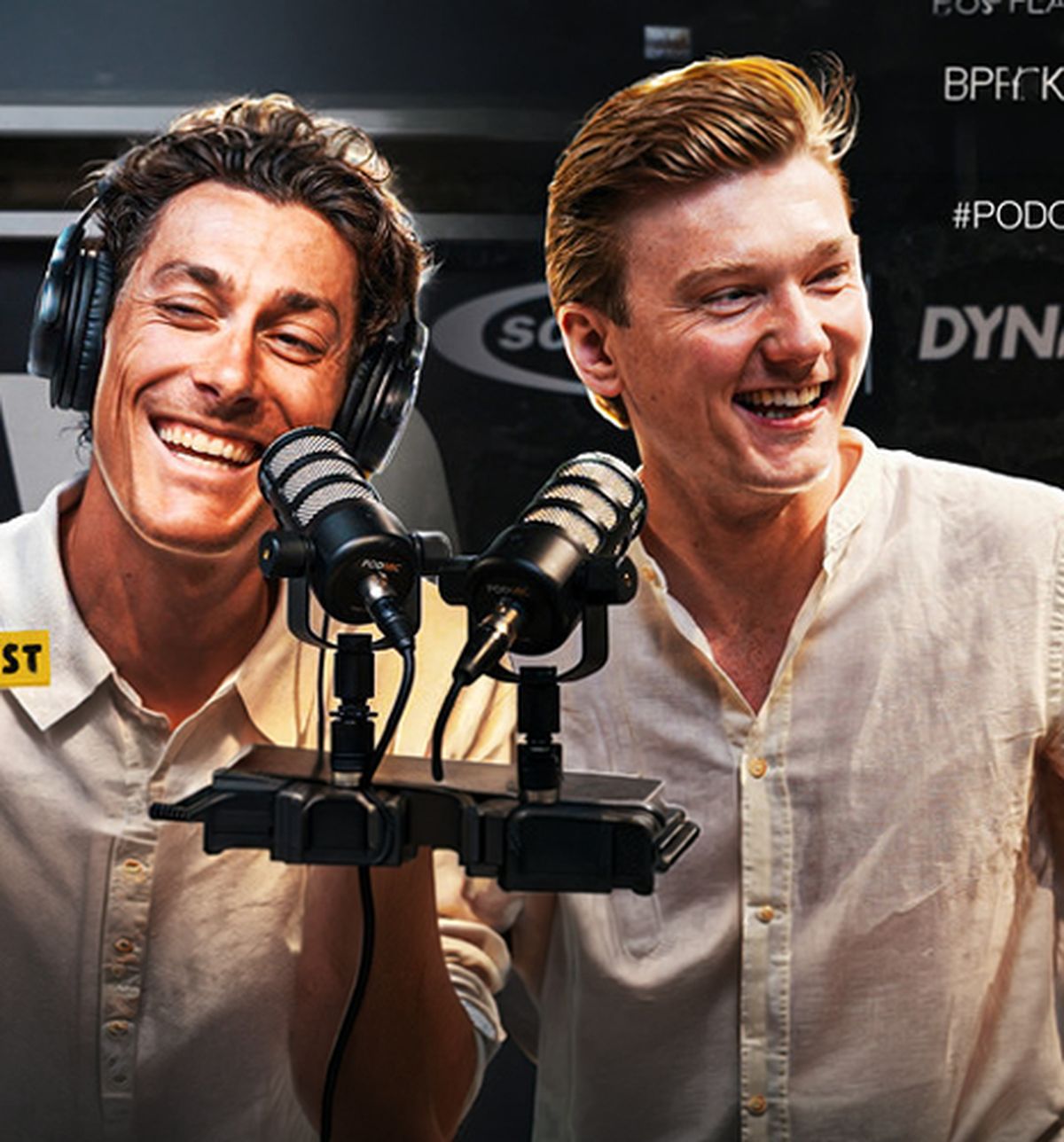 Two men in white shirts laughing into microphones, wearing headphones, in a studio setting.