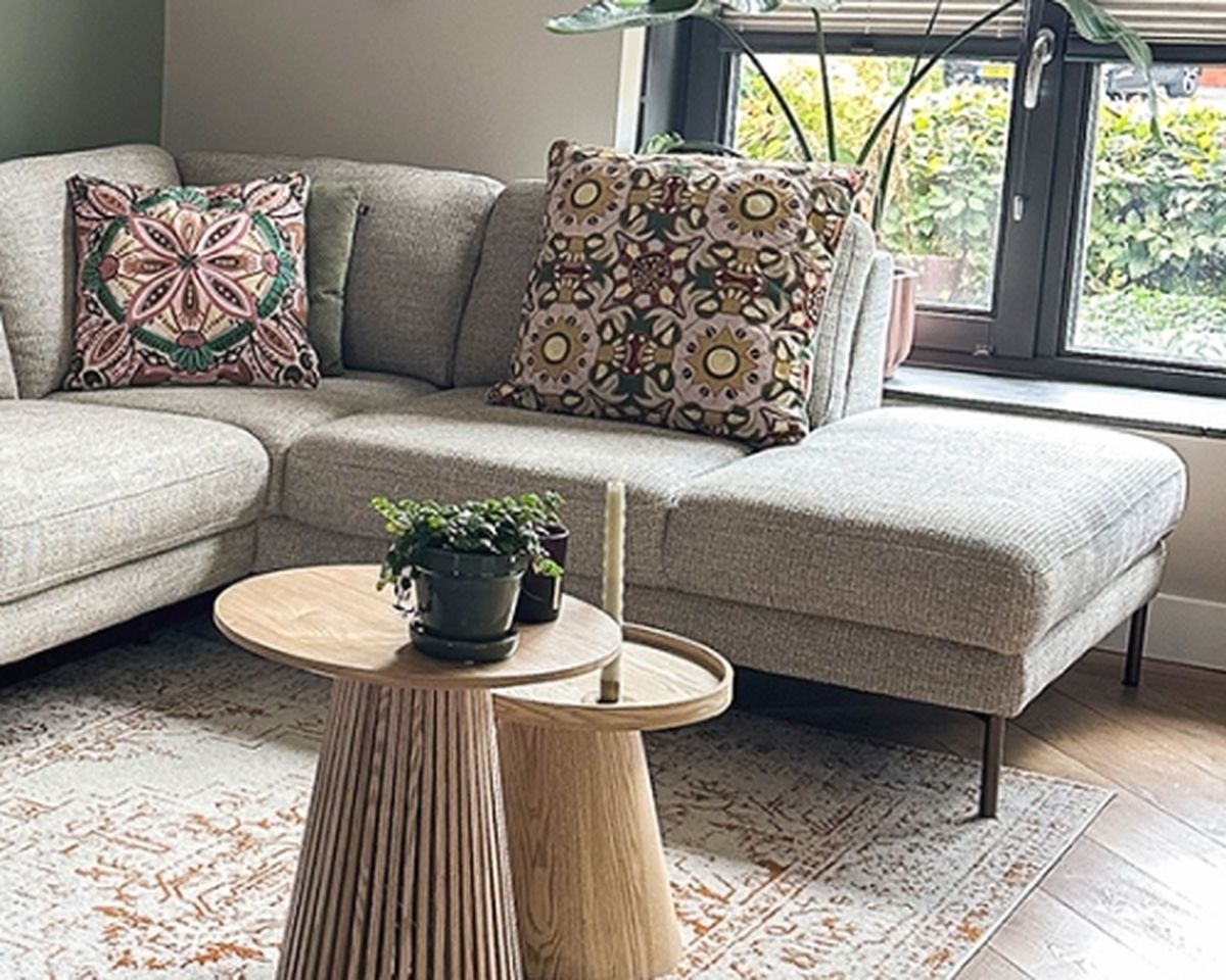 A modern living room with a gray sectional sofa featuring colorful patterned cushions, a round wooden coffee table, and a plant by the window.