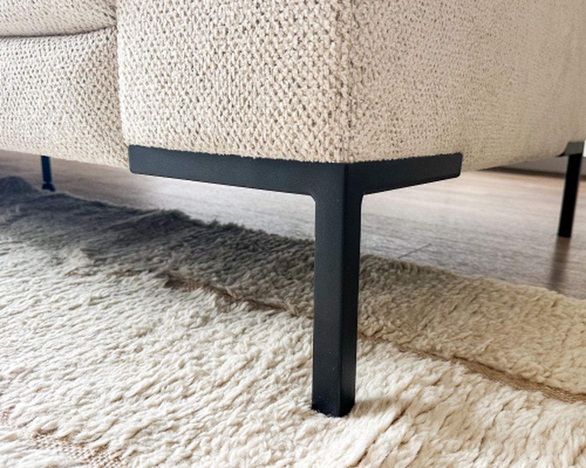 Close-up van de hoek van een beige bank met textuur en zwarte metalen poten, op een crèmekleurig geweven vloerkleed op een houten vloer.
