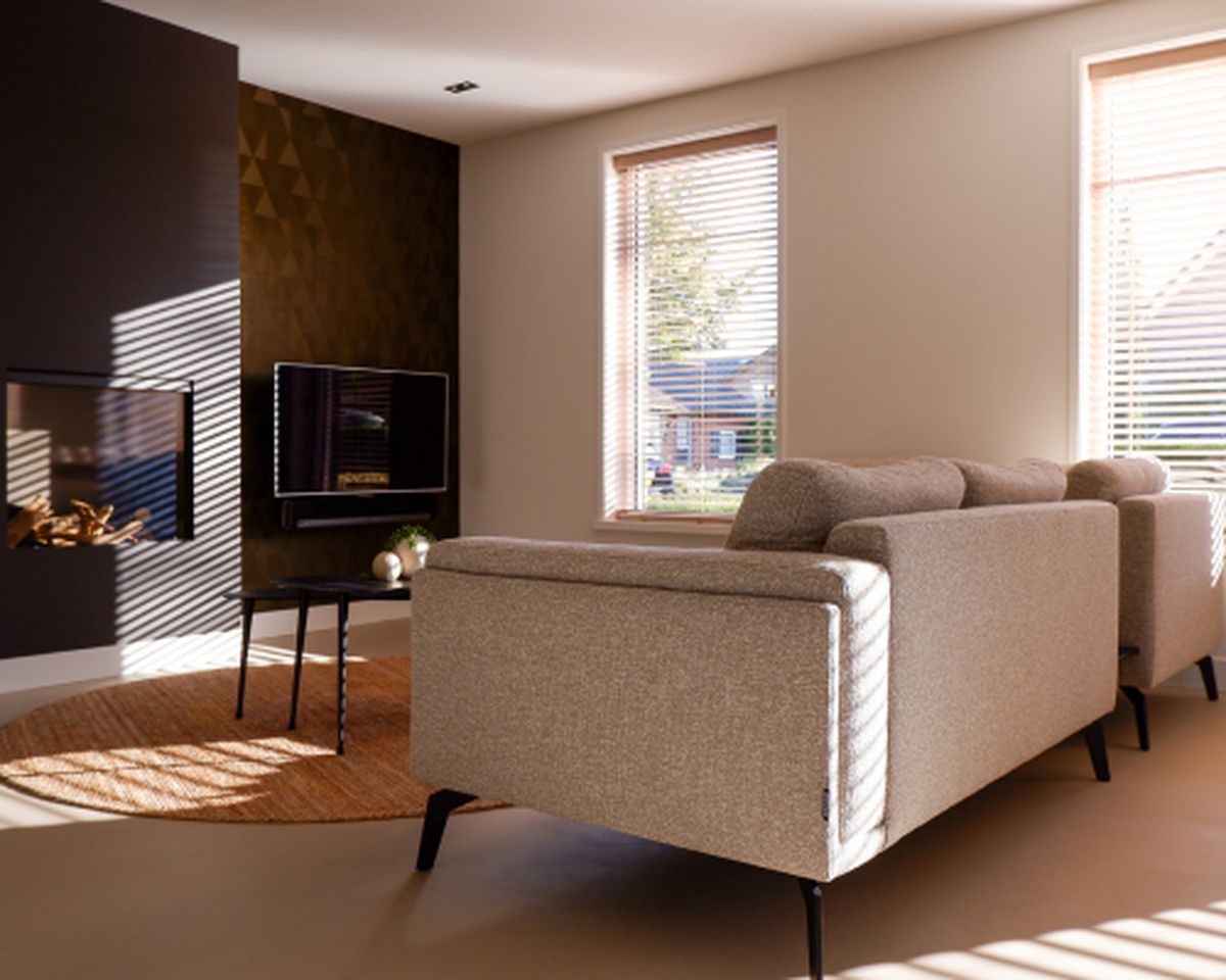 Modern ingerichte woonkamer met een grijze bank, een ronde tafel, een televisie en een open haard. Zonlicht valt door de jaloezieën naar binnen.
