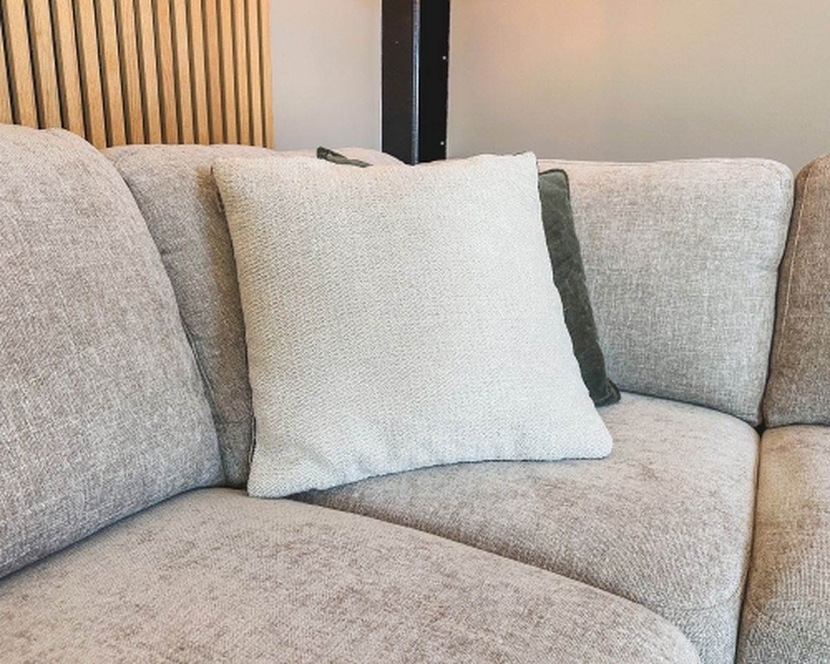 Beige fabric corner sofa with a textured cream pillow and a darker pillow in a cozy living room setting. Vertical wood panel detail on the wall.