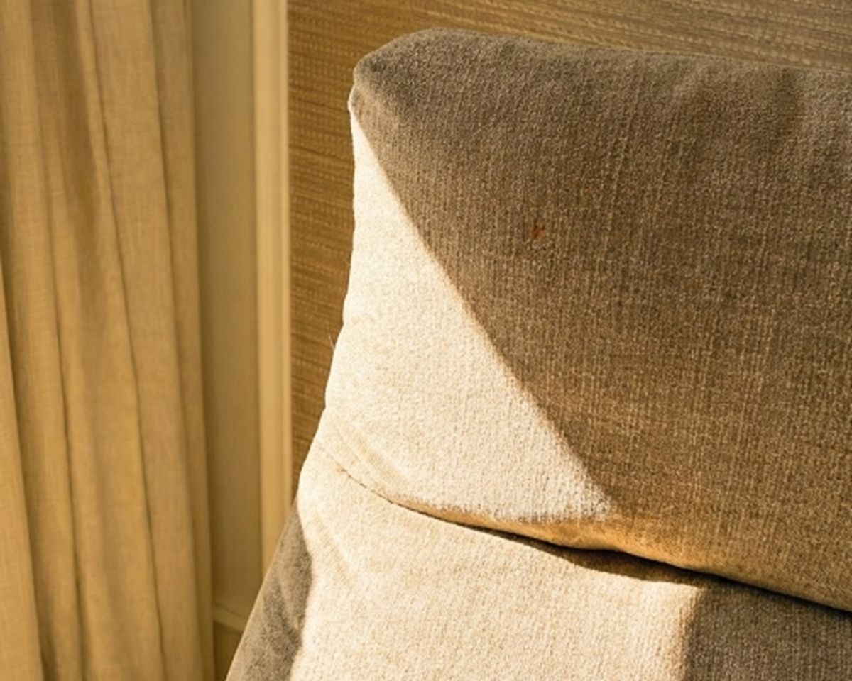 Close-up of a beige sofa backrest with sunlight casting shadows on it. Nearby is a curtain in the background, creating a cozy and warm ambiance.