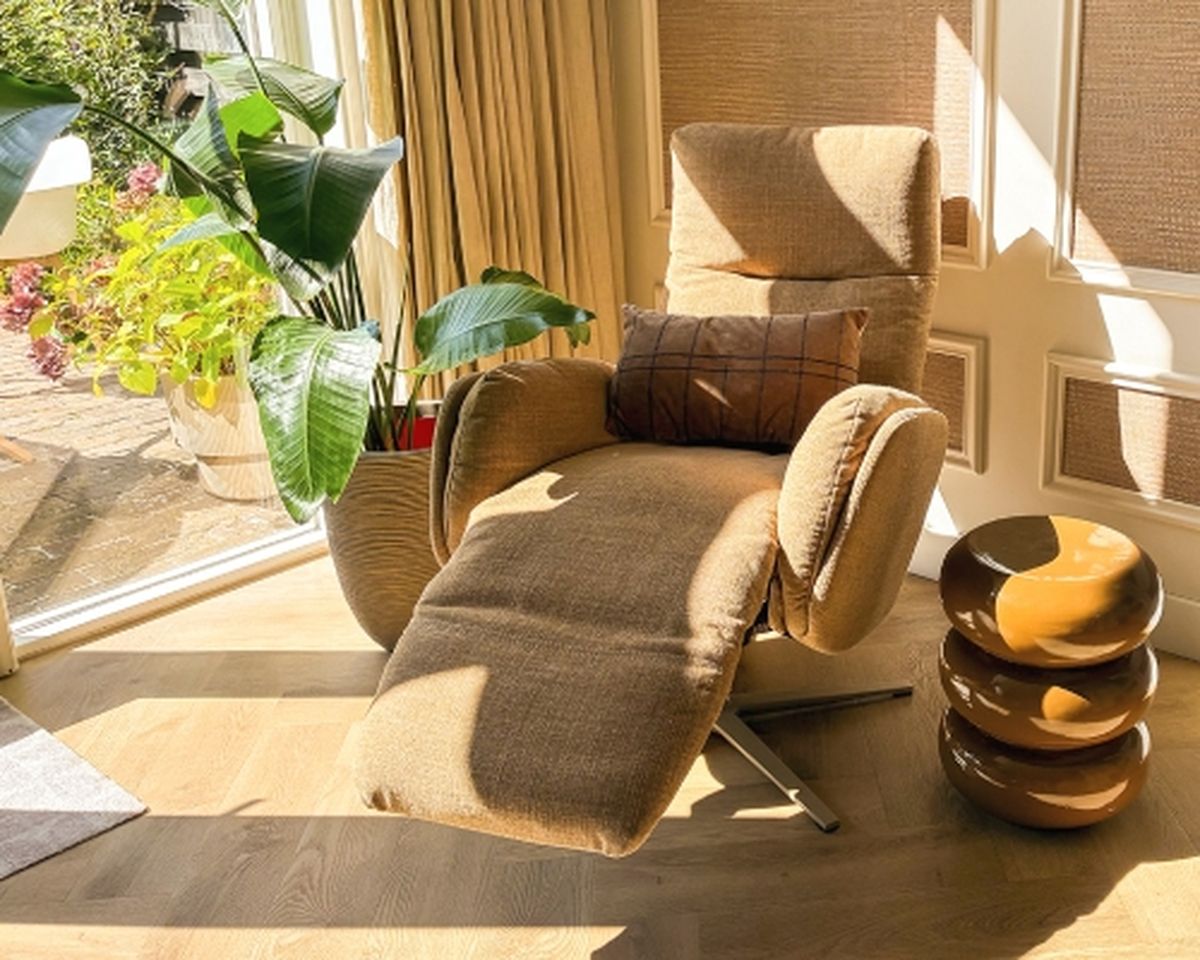 A light brown reclining chair with a pillow next to a large potted plant and stacked round tables in a sunlit room.