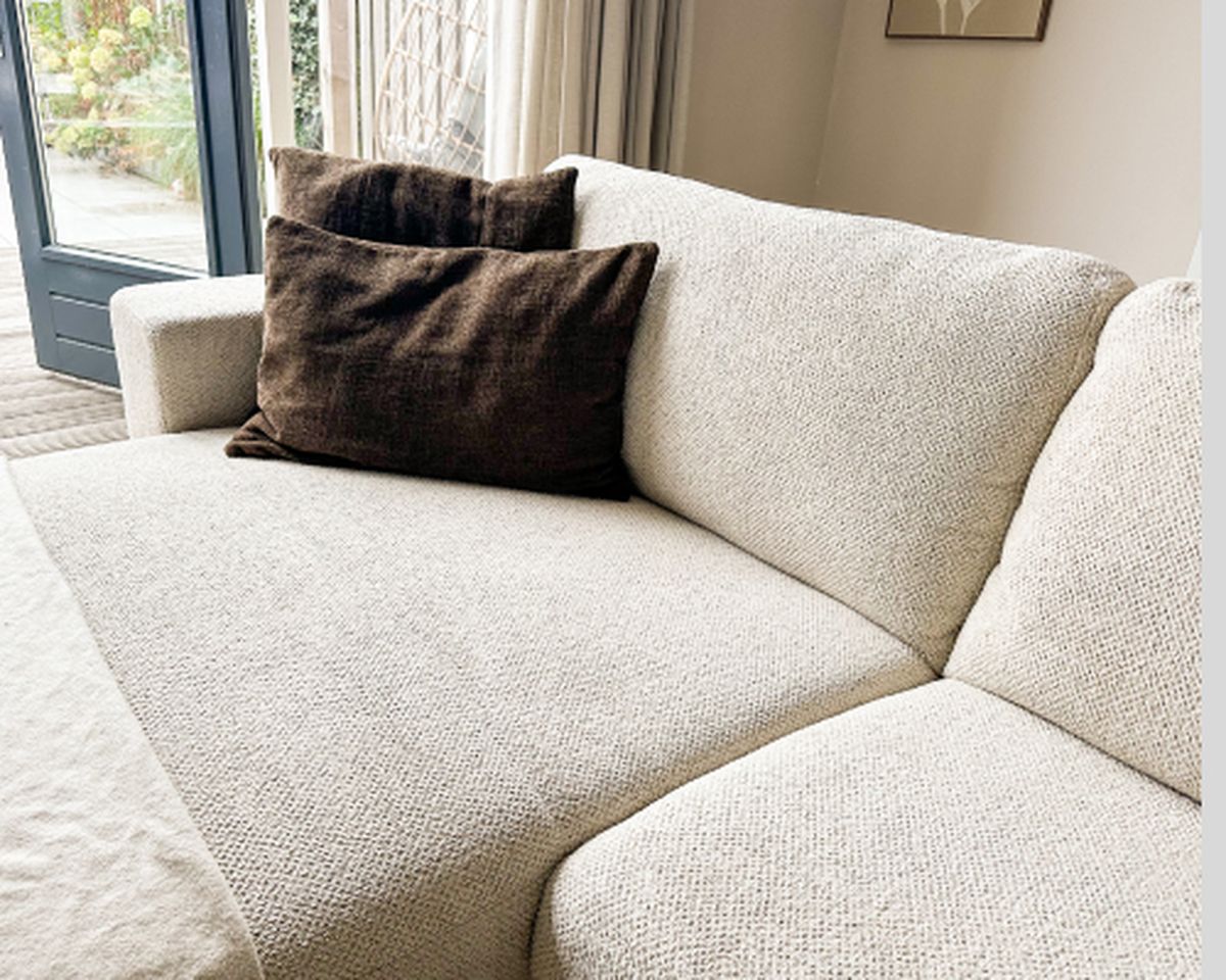 Lichtbeige hoekbank met twee bruine kussens in een woonkamer, bij een raam met vitrage.