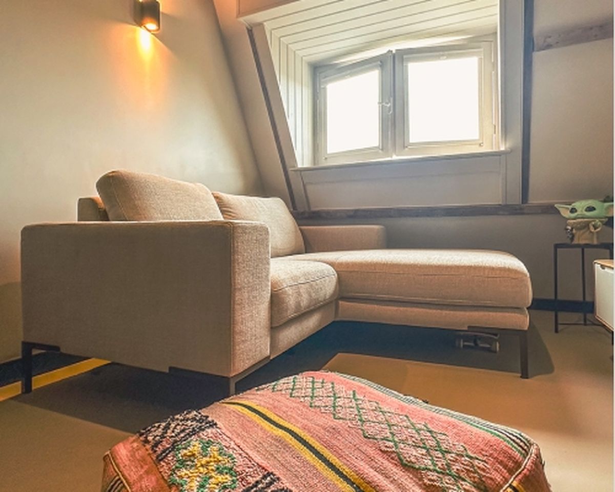 Cozy attic room with a beige sectional sofa, a side table with a decorative object, a colorful patterned rug, and a small window letting in light.