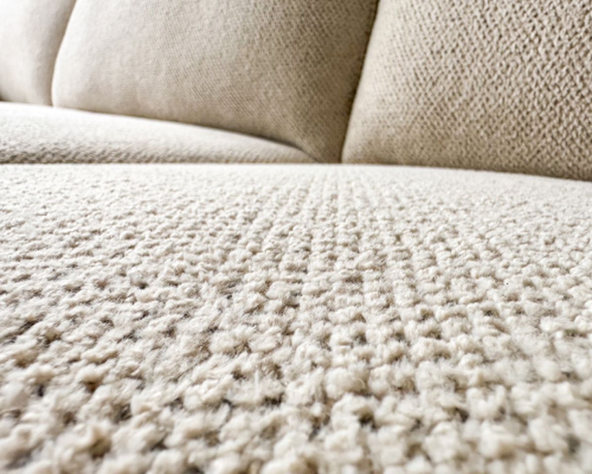 Close-up van een beige stof met textuur, waarop een verfijnd geweven patroon en zachte bekleding te zien zijn; onderdeel van een bank.