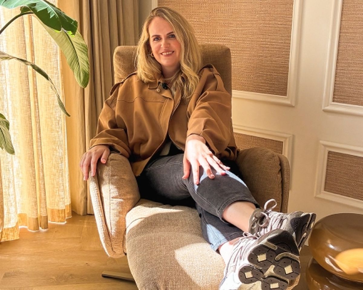 A woman with long blonde hair, wearing a brown jacket, black jeans, and sneakers, sits relaxed in a chair near a window with a potted plant.