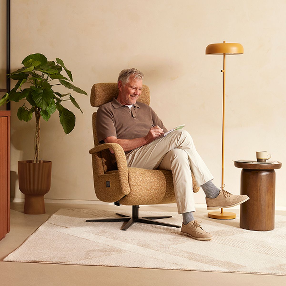 A man sits in a cozy chair, writing on a notepad, beside a potted plant and a floor lamp in a warmly lit room.