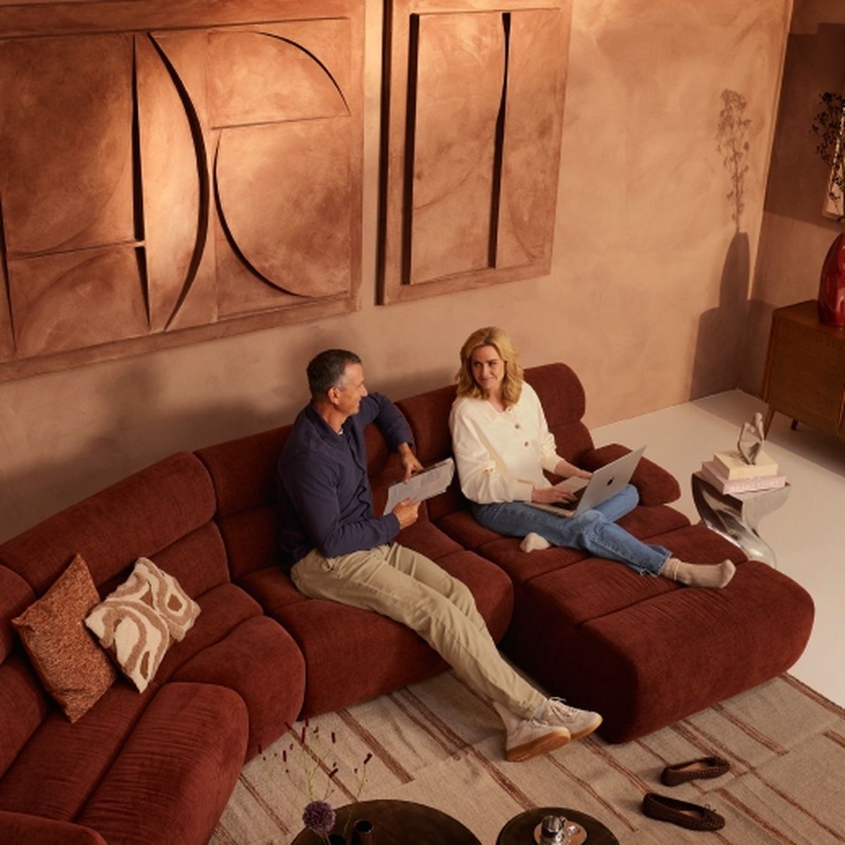 A man and a woman sit on a brown sectional sofa, discussing papers and using a laptop, with abstract art on the wall behind them.