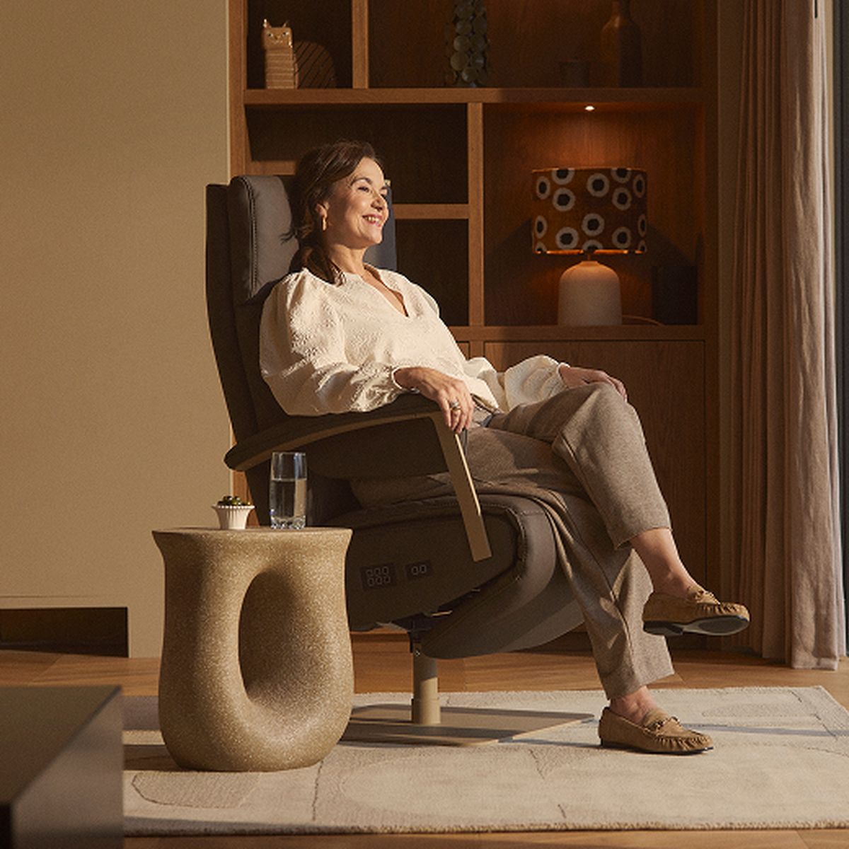 A woman relaxes in a reclining chair, smiling next to a modern side table with a glass of water, in a cozy room with warm lighting and shelves.