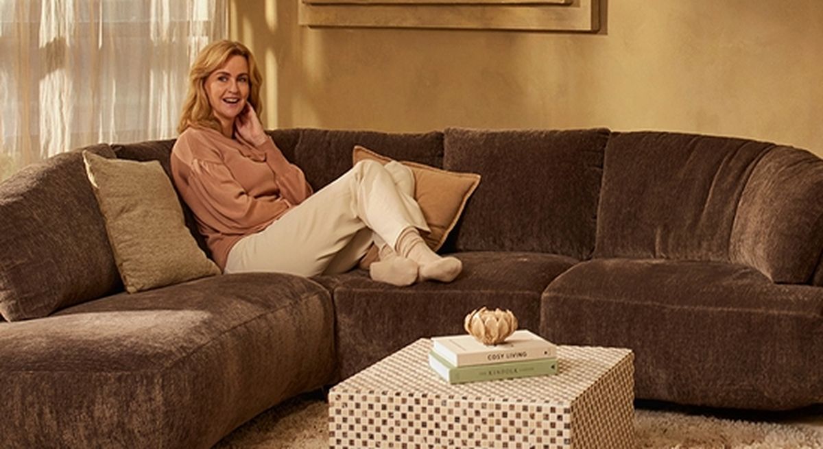 A woman lounges on a large, brown sectional sofa with pillows, smiling warmly. A checkered coffee table holds books and a decorative object.