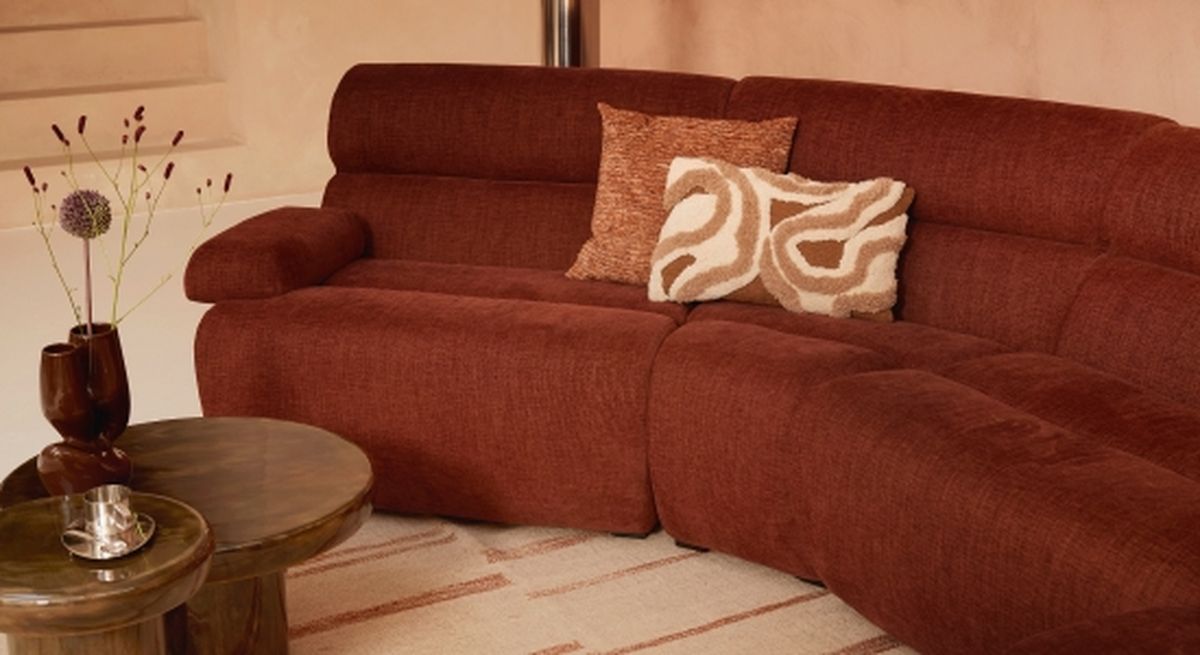 Bruine bank met kussens met textuur, naast een houten salontafel en een vaas met bloemen, geplaatst op een lichtgekleurd tapijt in een knusse kamer.