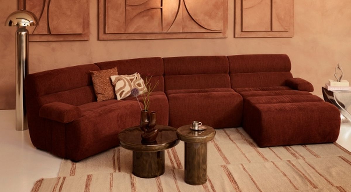 A cozy living room with a brown sectional sofa, patterned cushions, round wooden tables, and modern wall art in a warm, earthy color palette.