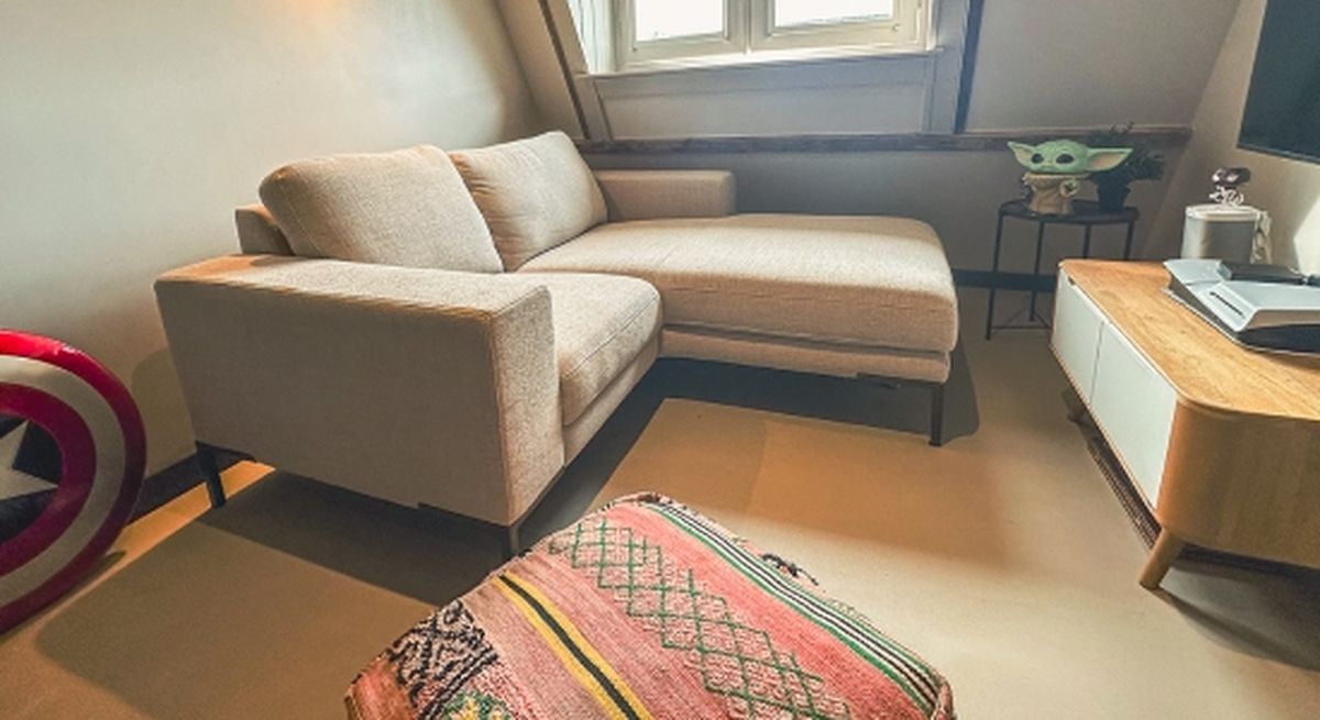 Cozy attic living space with a beige sectional sofa, patterned ottoman, and a small side table holding decorative items, lit by a window.