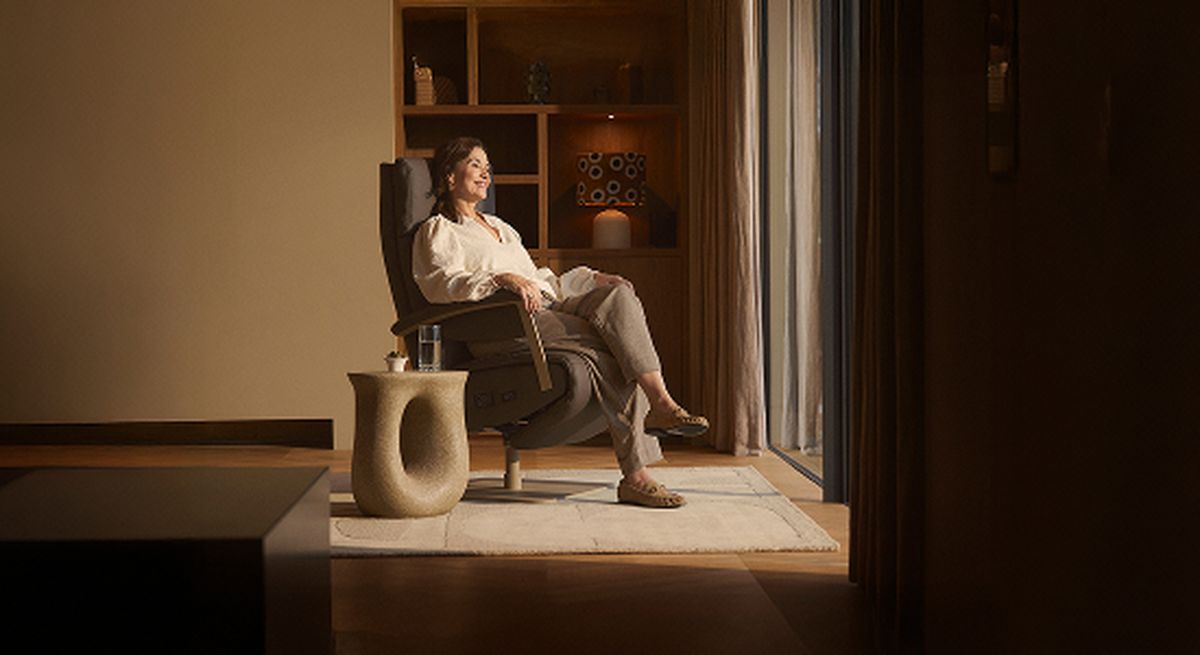 A woman sits relaxed in a comfortable chair by a window, bathed in warm light, with a side table nearby, in a cozy, modern room.