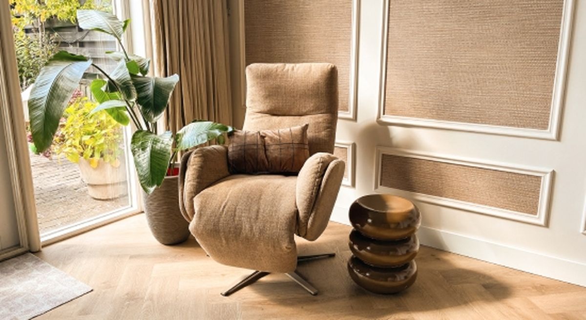 A cozy beige recliner with a patterned cushion beside a tall green plant, near a sunny window and a stack of round, glossy brown stools.