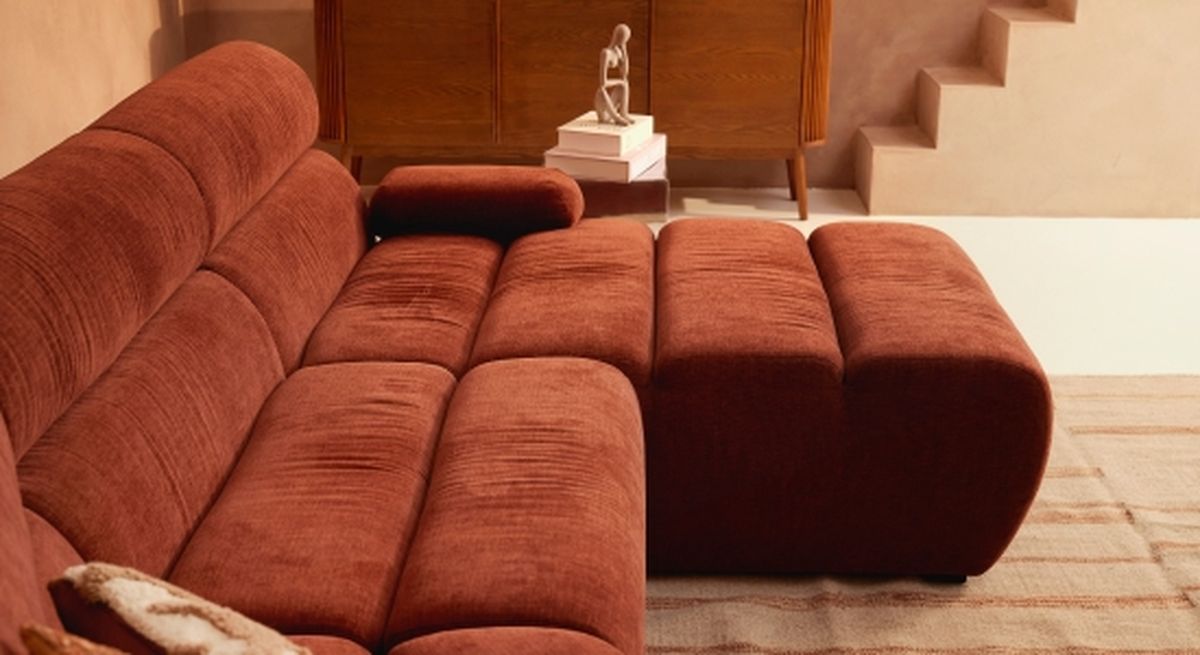 Roestkleurige hoekbank met zachte bekleding in een knusse woonkamer. Op de achtergrond staan ​​een houten kast en een decoratief beeldje.