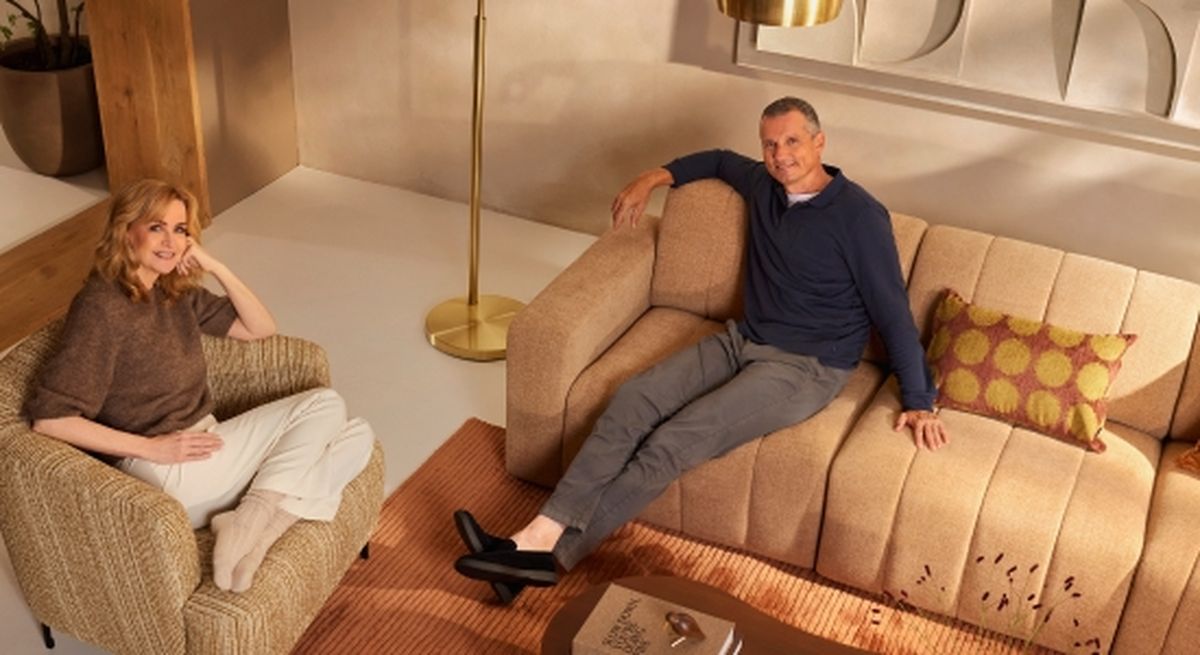 A man and woman sitting comfortably in a well-lit living room. The woman smiles from an armchair, and the man relaxes on a sofa with a decorative cushion.