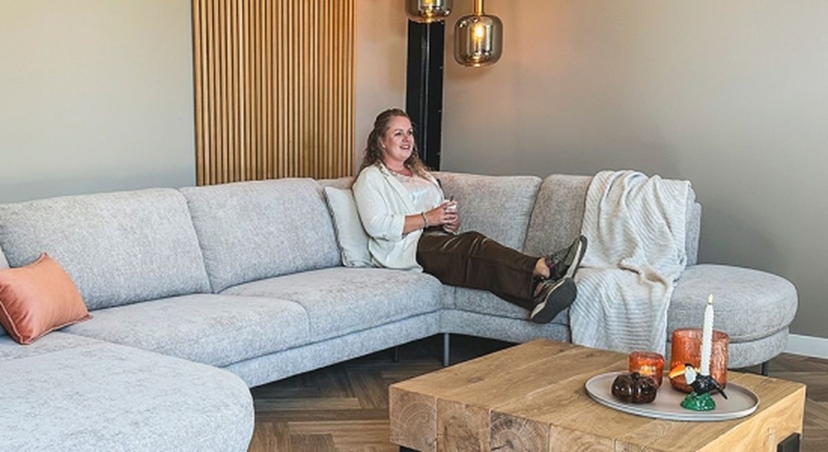 A person relaxes on a gray sectional sofa, holding a cup, with cozy decor and a wooden coffee table in a stylish living room.