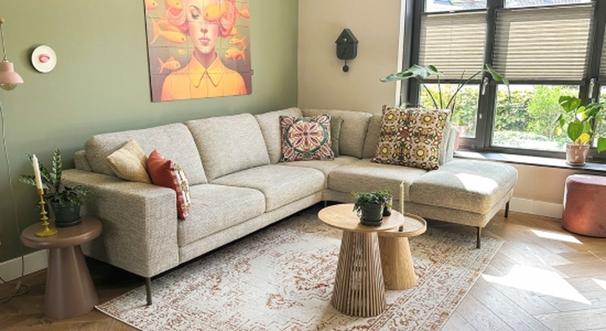 A cozy living room with a gray sectional sofa adorned with patterned cushions, wooden tables, indoor plants, and a colorful wall art on a green wall.
