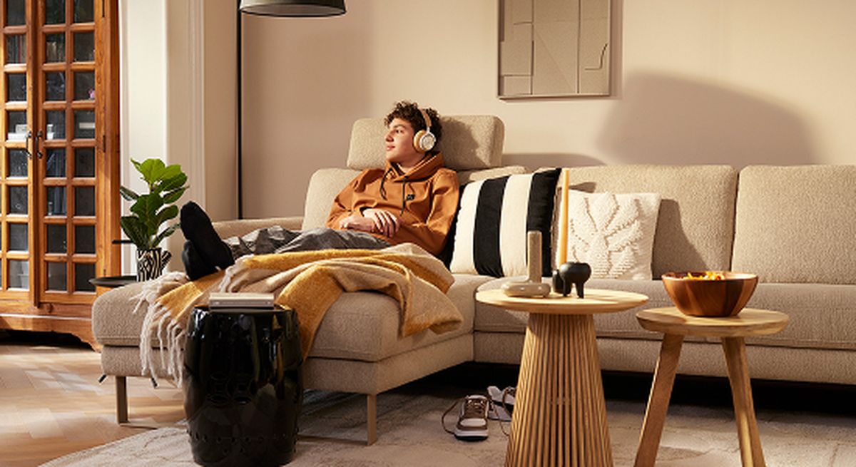 A person relaxing on a beige couch with headphones, surrounded by modern decor, including a plant, tables, and a bowl of snacks.