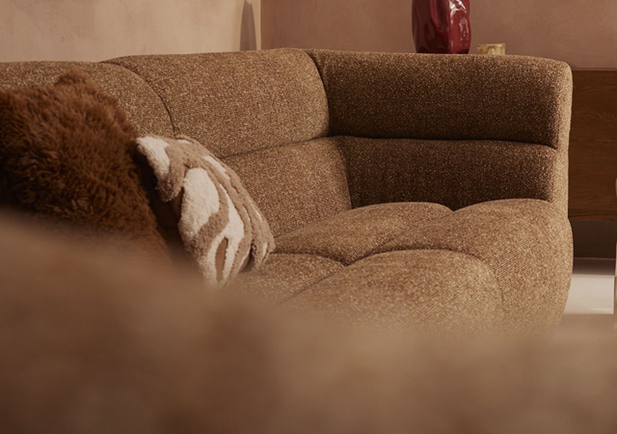 A cozy brown textured sofa with plush throw pillows, set against a warm-toned wall. A decorative red vase is partially visible in the background.
