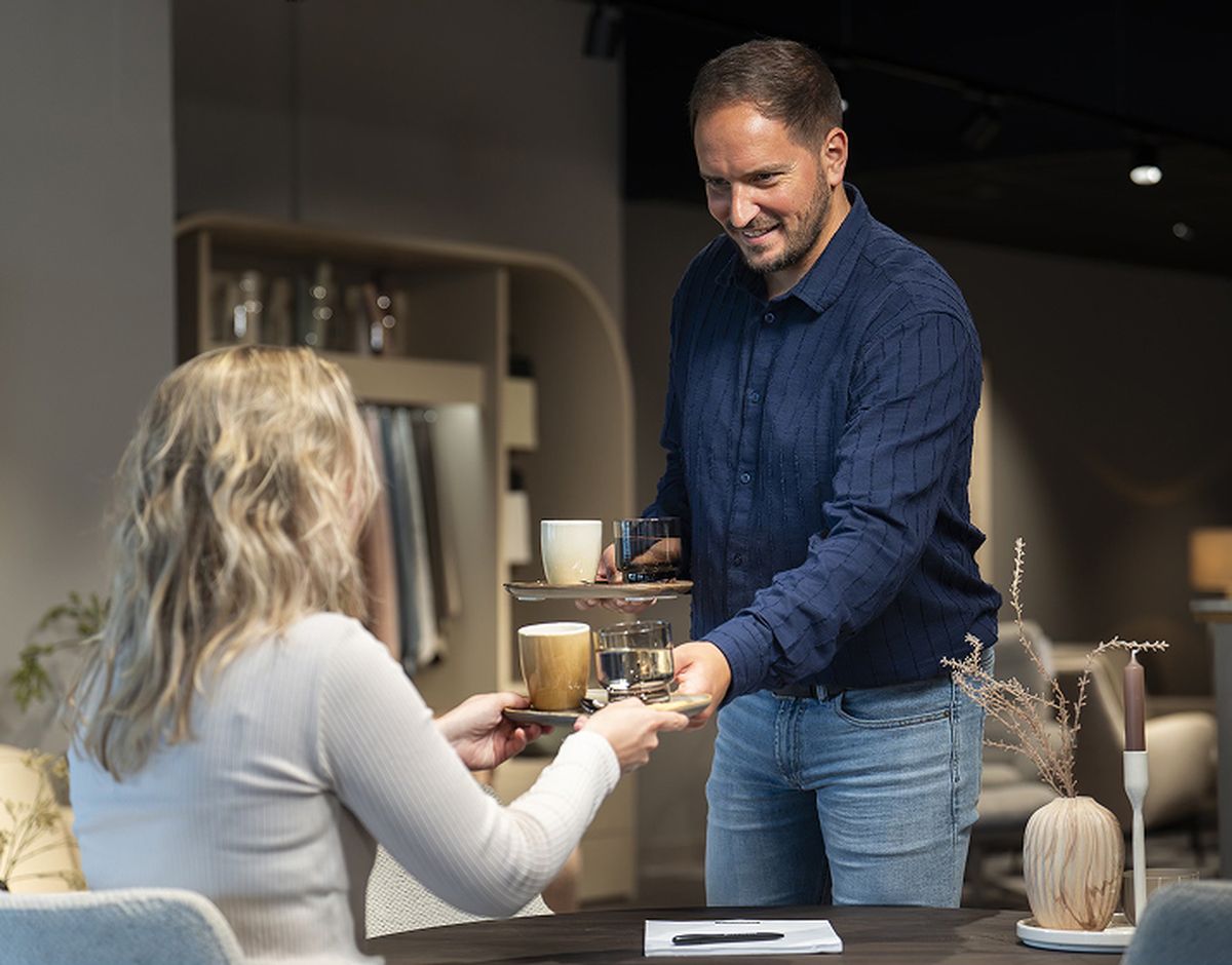 Een man in een blauwe blouse serveert drankjes aan een zittende vrouw met blond haar in een kantoorachtige omgeving.