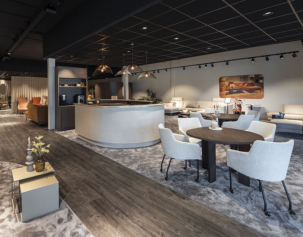 Moderne woonkamer showroom met comfortabele banken, ronde tafel met stoelen, warme verlichting en decoratieve accessoires op houten vloer en tapijt.