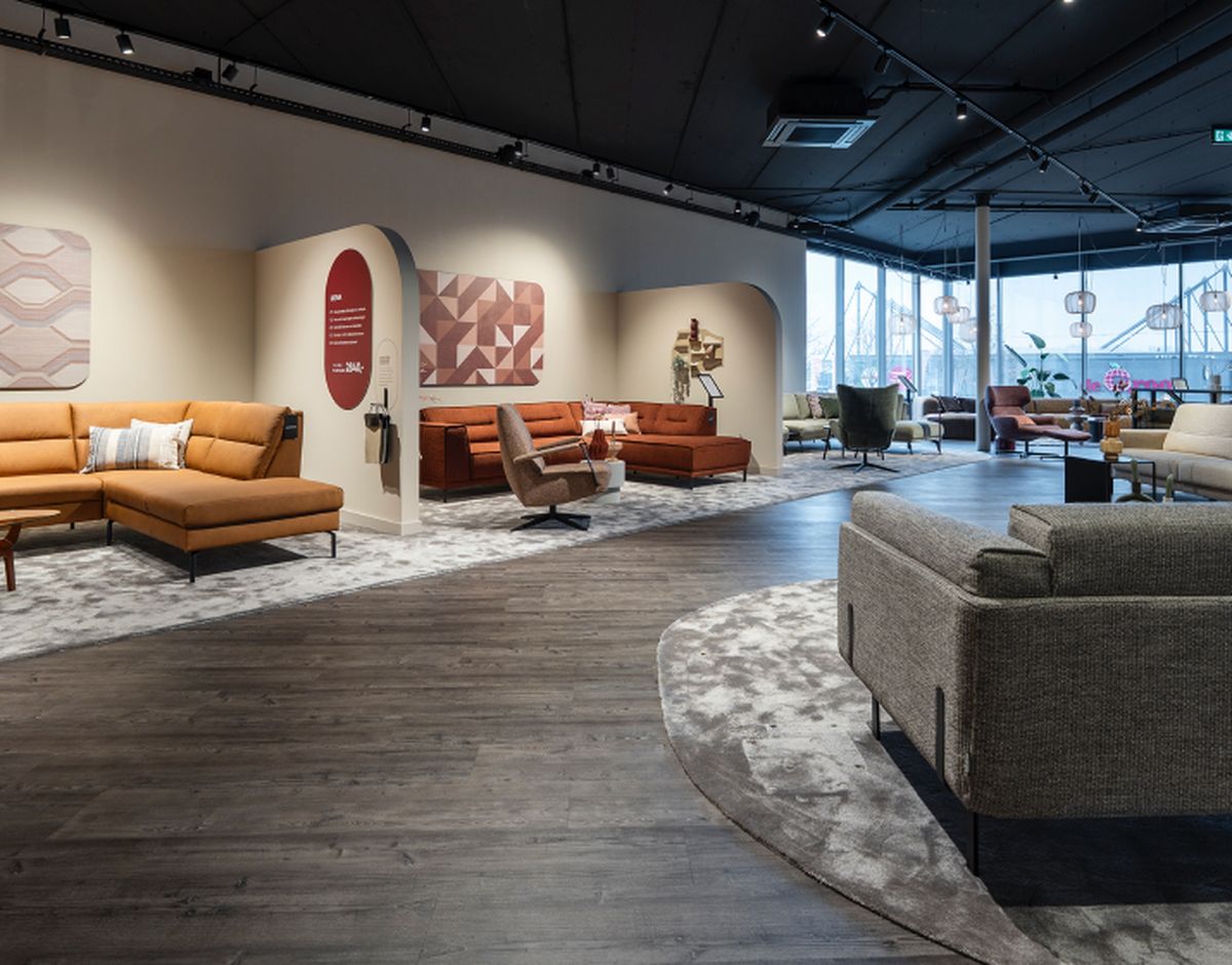 Ruime meubelwinkel met moderne banken, fauteuils en decoratieve wanden. Grote ramen laten veel licht binnen. Houten vloeren en tapijten.