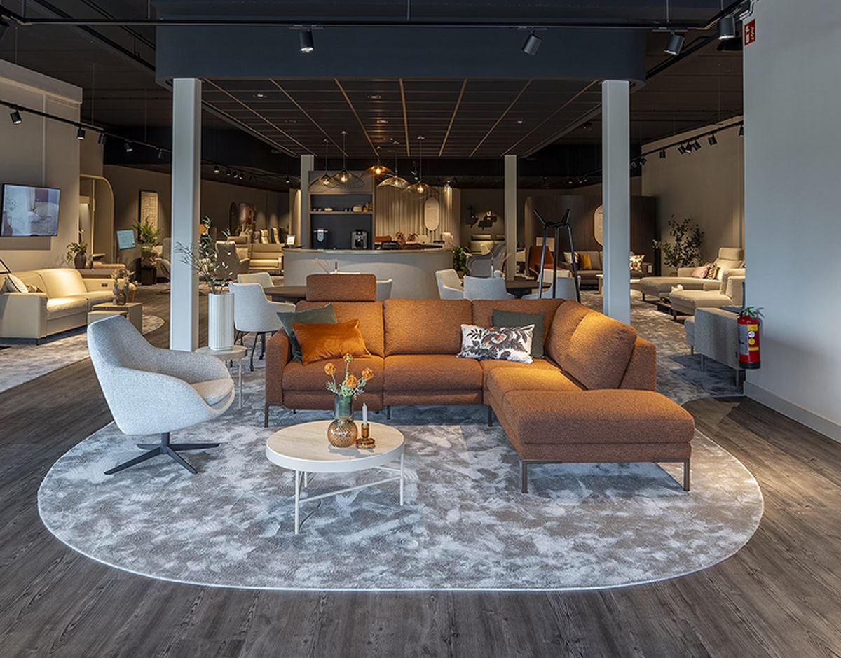 Moderne woonkameropstelling met oranje hoekbank, grijze fauteuil, wit salontafeltje op een rond tapijt, donkere houten vloer en sfeervolle verlichting.