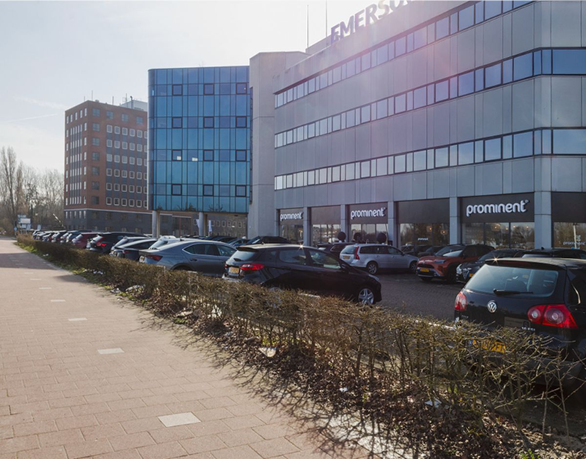 Parkeerplaats met auto's voor een modern kantoorgebouw met donkere glazen ramen. Zonnige dag met een rij kale struiken langs het trottoir.