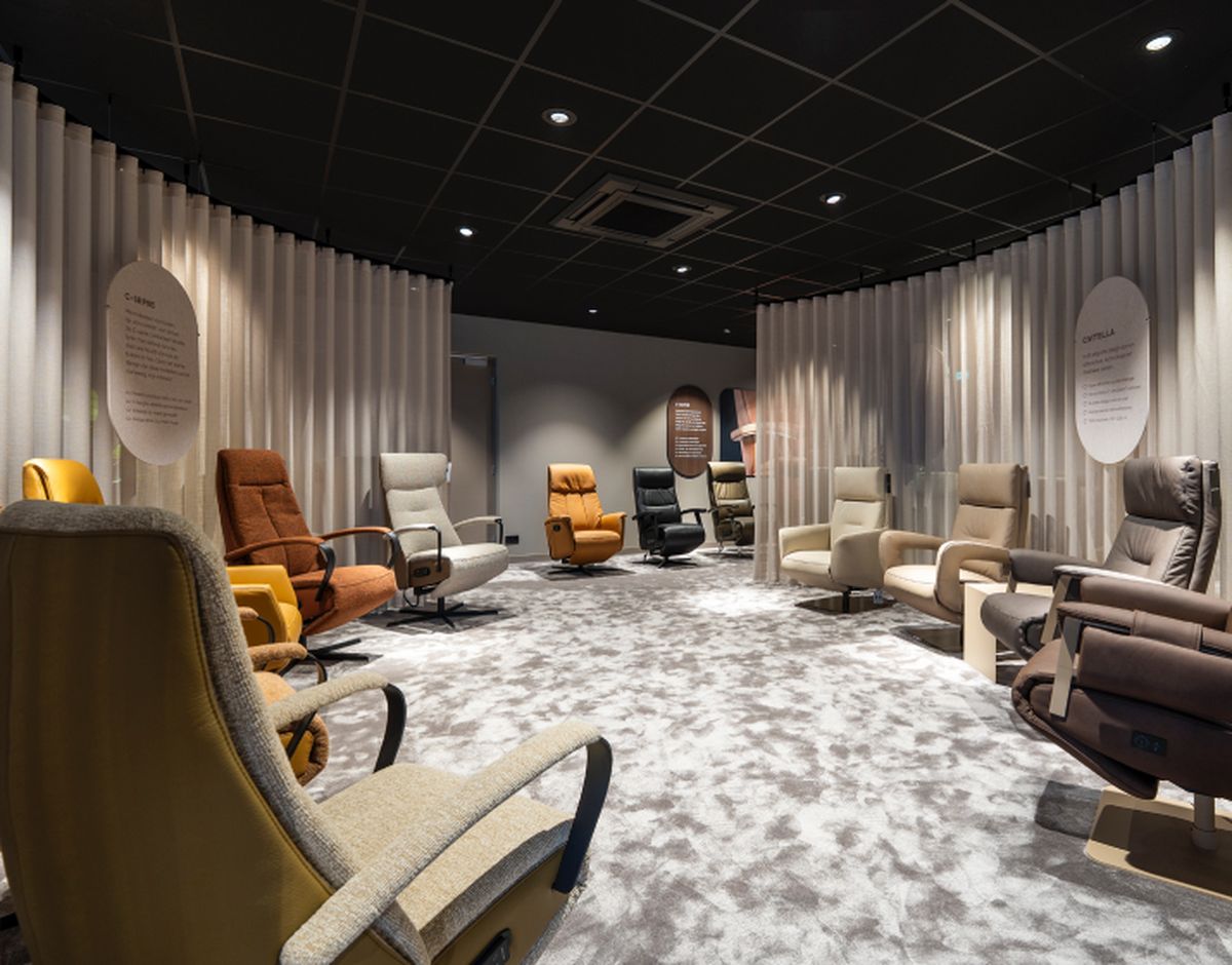 Een showroom met een cirkelopstelling van comfortabele fauteuils in diverse kleuren en stijlen op een vloer met een grijs patroon, omgeven door gordijnen.