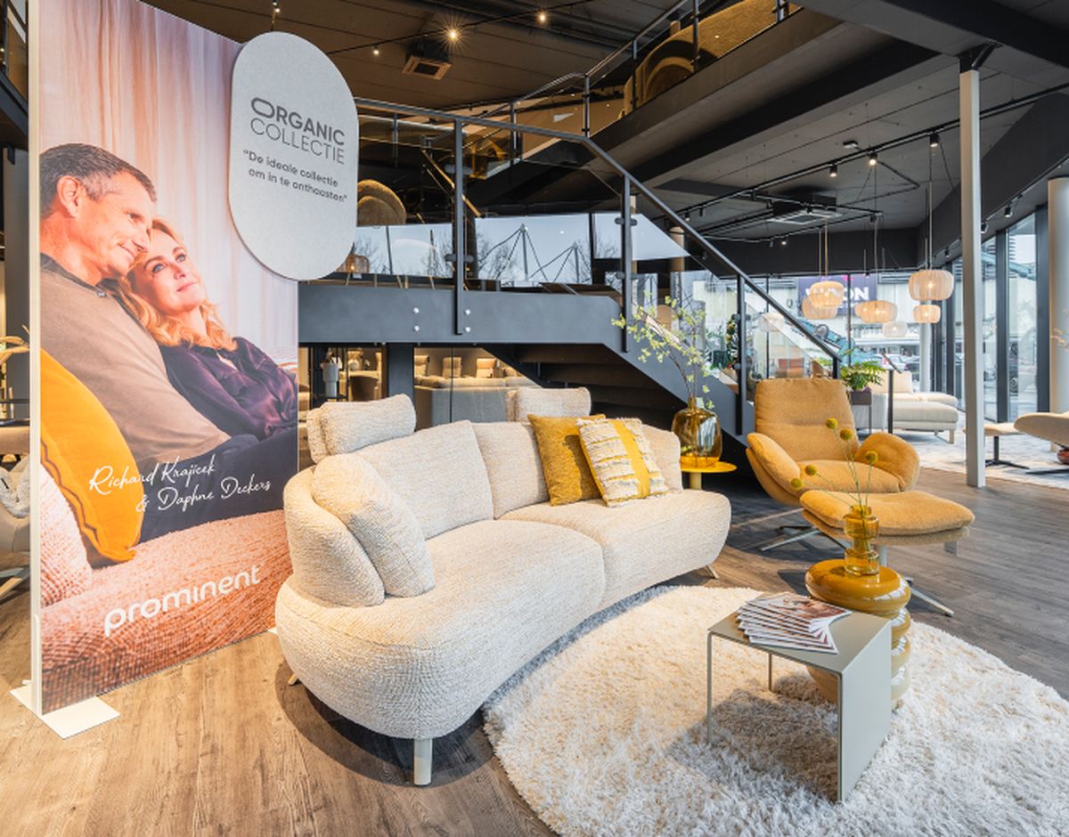 Moderne meubelwinkel met wit crèmekleurige bank, gele stoel, en een reclamebord. Lichte en open inrichting met houten vloer en stalen trap.