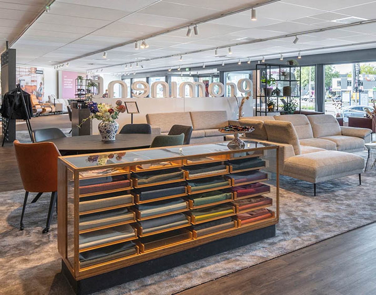 Moderne meubelwinkel met comfortabele banken, een tafel met stoelen en een toonbank met stoffenmonsters. Ruime, lichte sfeer met grote ramen.