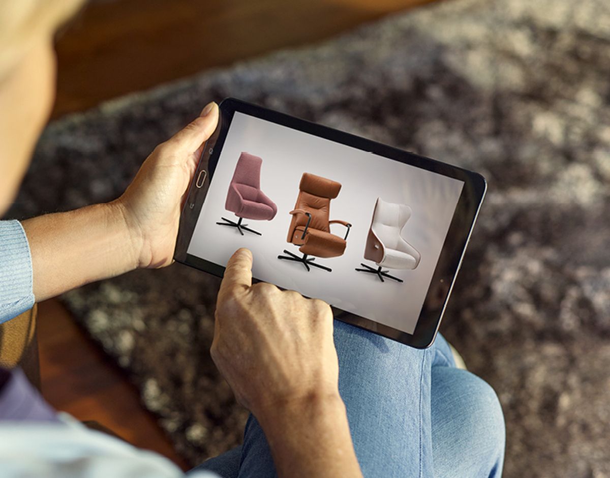 Persoon bekijkt op tablet drie moderne stoelen in verschillende kleuren, zittend op een vloerkleed.